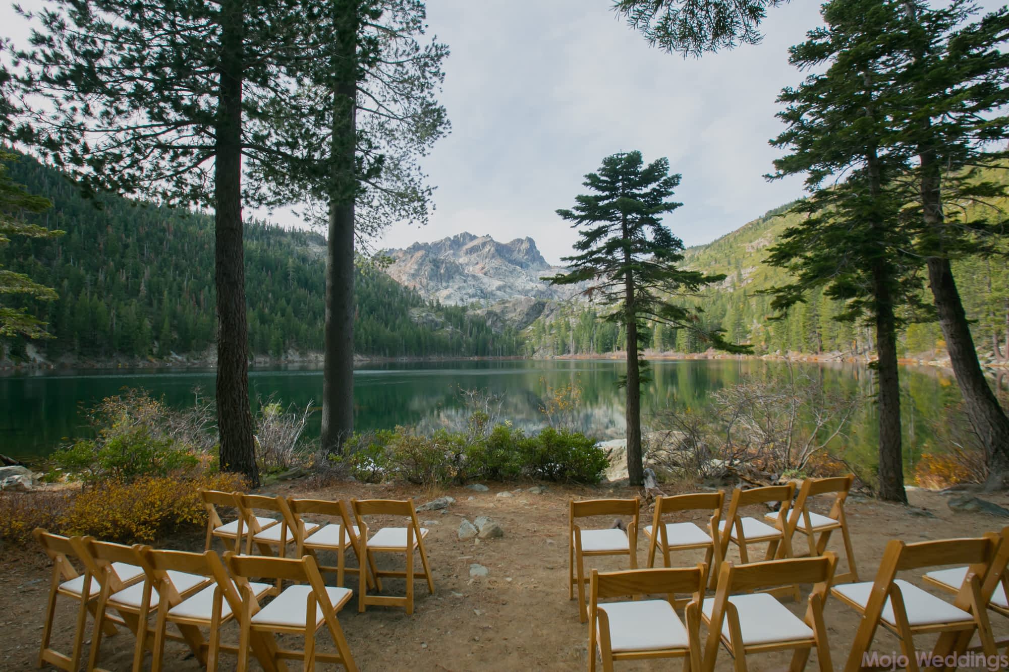 Chairs are set up for a wedding ceremony in front of a lake and pine trees.