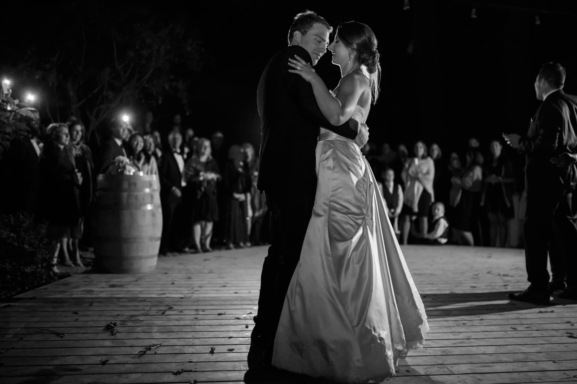 Black and white moody image of the bride and groom dancing with the dimly lit guests in the background.