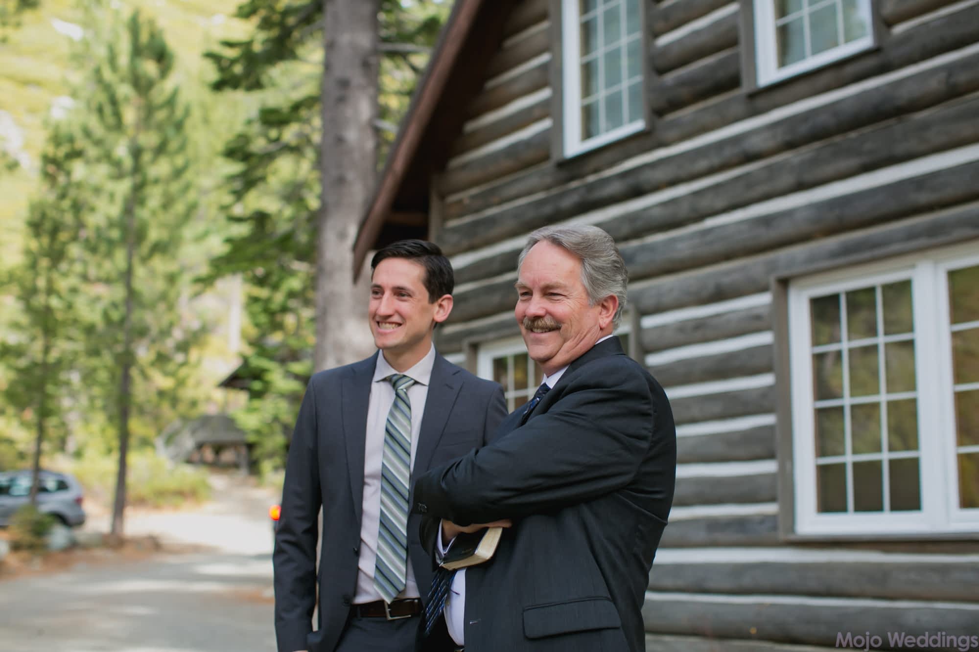 The groom and officiant smile before the ceremony with a log cabin in the background.