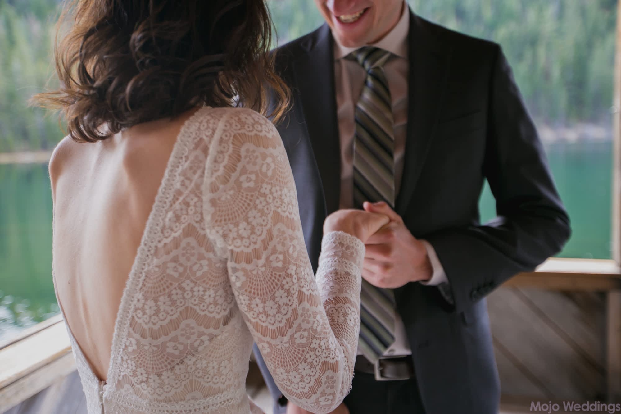 A close up of the brides lacy dress leads as the groom holds her hand.