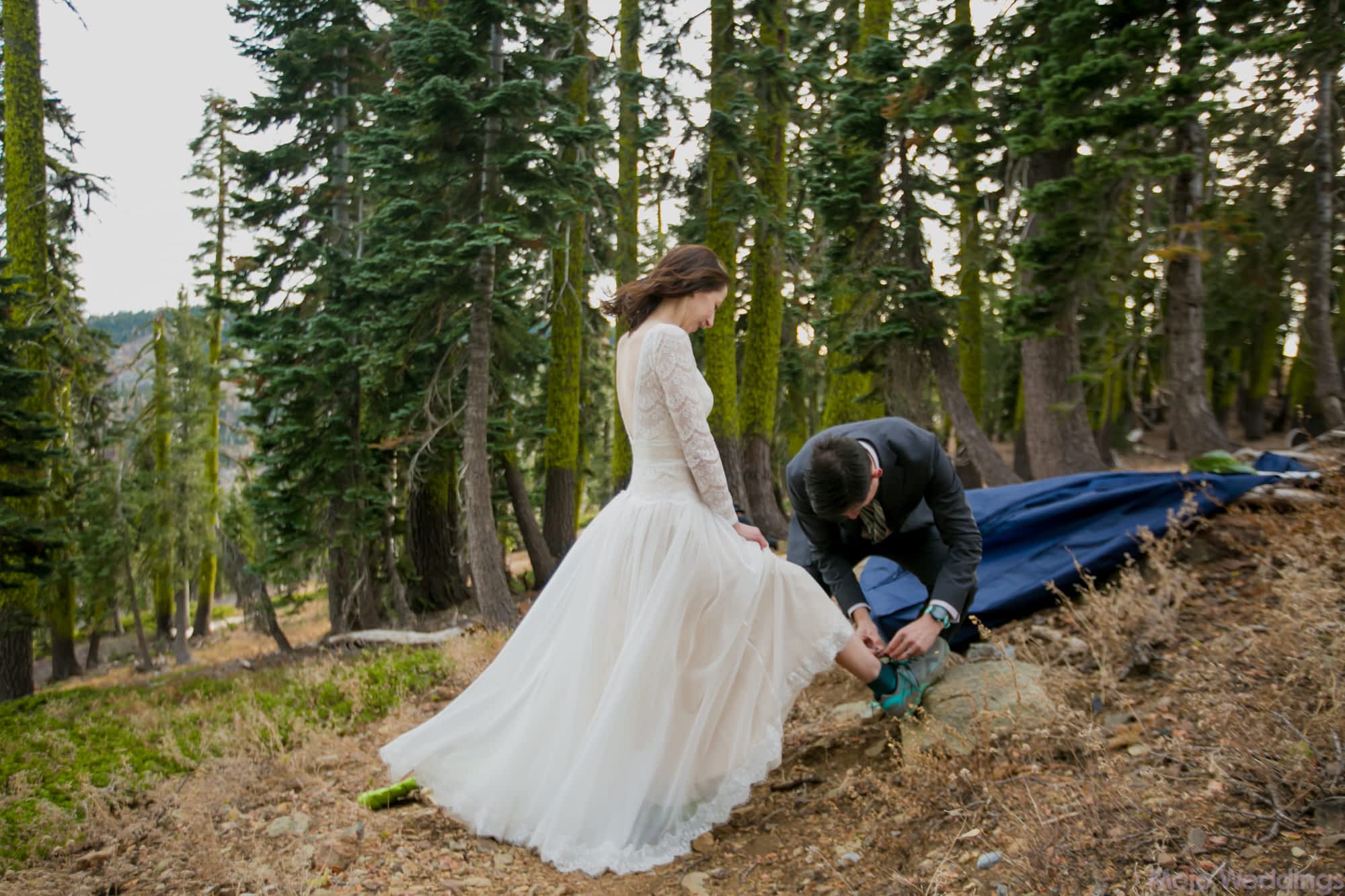 Her white dress fans out white he helps lace up her hiking shoes.