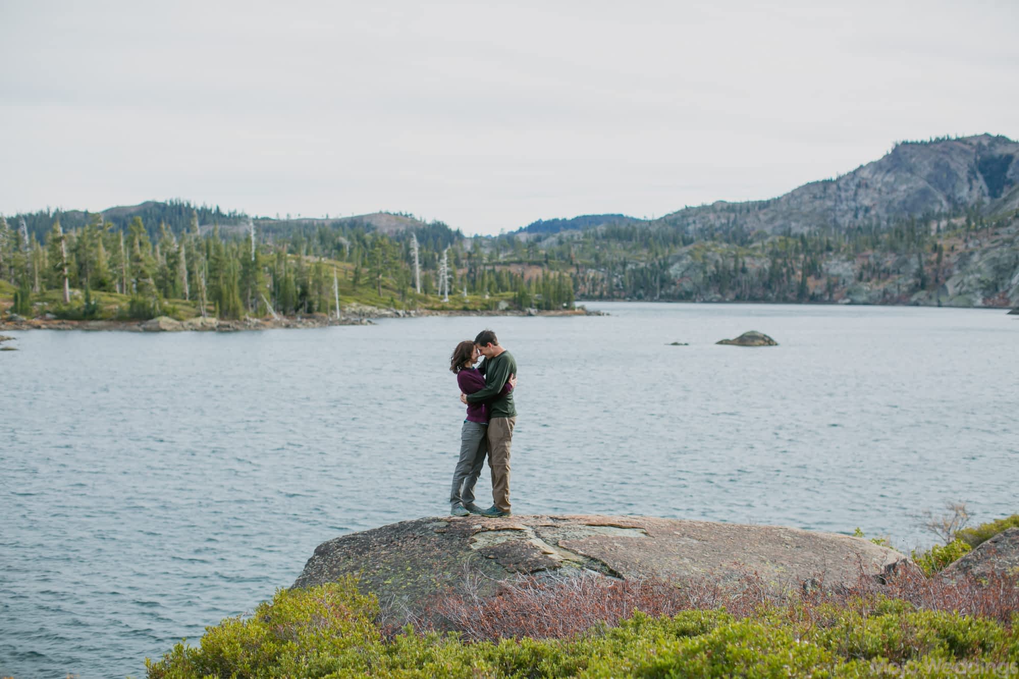 The bride and groom, dressed in hiking clothes, embrace right next to a lake.